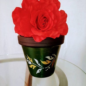 May include: A single red rose in a green terracotta pot with a white and gold floral design. The pot is sitting on a glass table.