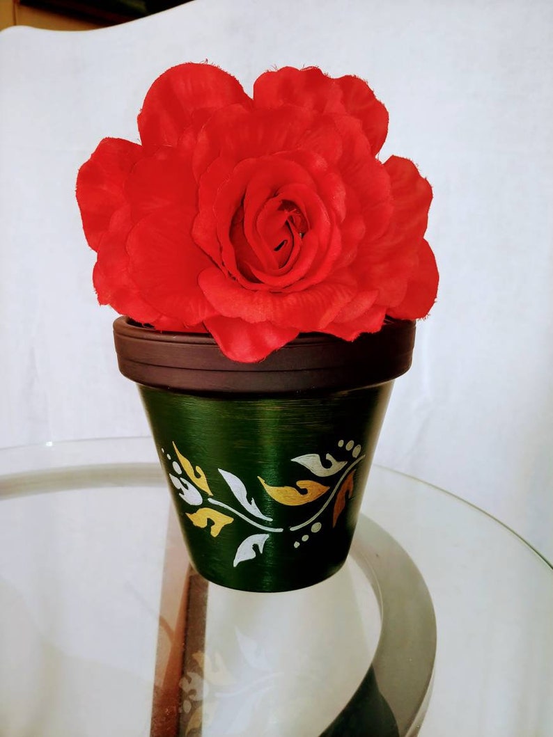 May include: A single red rose in a green terracotta pot with a white and gold floral design. The pot is sitting on a glass table.