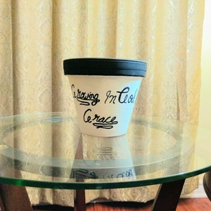 May include: White ceramic flower pot with a black rim. The pot has black lettering that reads "Growing in God's Grace".