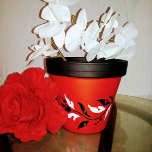 May include: A red terracotta flower pot with a black rim and white floral design. The pot is decorated with black and white dots. White orchid flowers are in the pot.