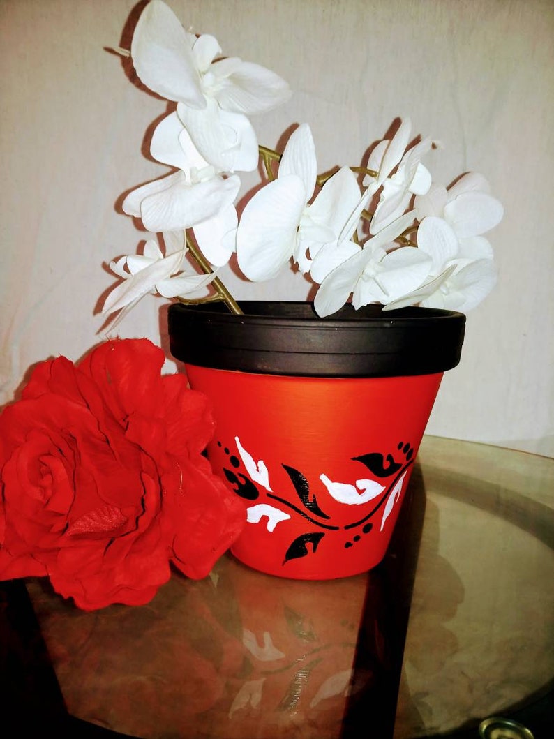 May include: A red terracotta flower pot with a black rim and white floral design. The pot is decorated with black and white dots. White orchid flowers are in the pot.