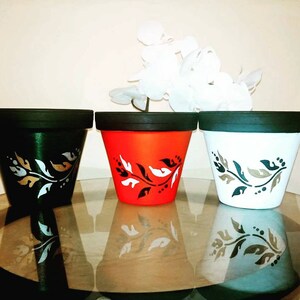 May include: Three terracotta flower pots with black rims, each painted in a different colour: green, orange, and white. Each pot has a black and gold floral design.