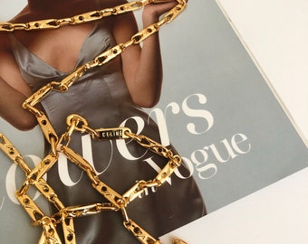 Op de afbeelding: Een gouden ketting met een rechthoekig label met de tekst "CELINE". De ketting is gemaakt van gekoppelde rechthoekige stukken met een textuur.