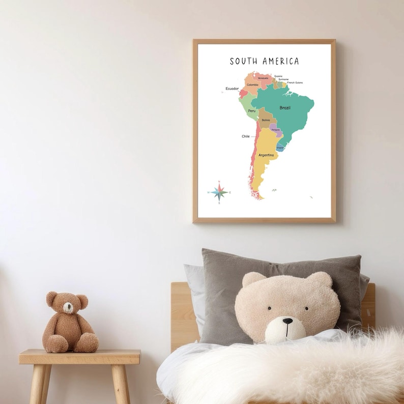 May include: A colorful map of South America with the countries labeled. The map is framed in a light wood frame and is hanging on a white wall. A teddy bear is sitting on a wooden stool in the foreground.