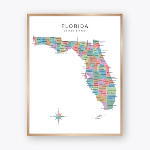 May include: A framed wall art print featuring a colorful map of Florida, United States. The map displays individual counties in various pastel shades with black text labels. A multi-colored compass rose is positioned in the lower left corner.