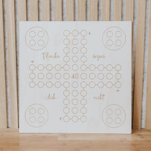 Könnte beinhalten: Ein Holzbrettspiel mit geätzten Linien und Kreisen. Das Brett zeigt die Wörter "Monika", "ärgert", "dich" und "nicht" sowie die Zahl "40". Vier Sätze von vier Kreisen befinden sich in jeder Ecke.