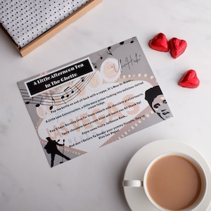 May include: A grey card with a musical note design and the text "A Little Afternoon Tea In The Chetto" and "Viva Las Vegas!"  The card also features a black and white illustration of a person singing into a microphone.  There are three red heart-shaped chocolates on a white surface.