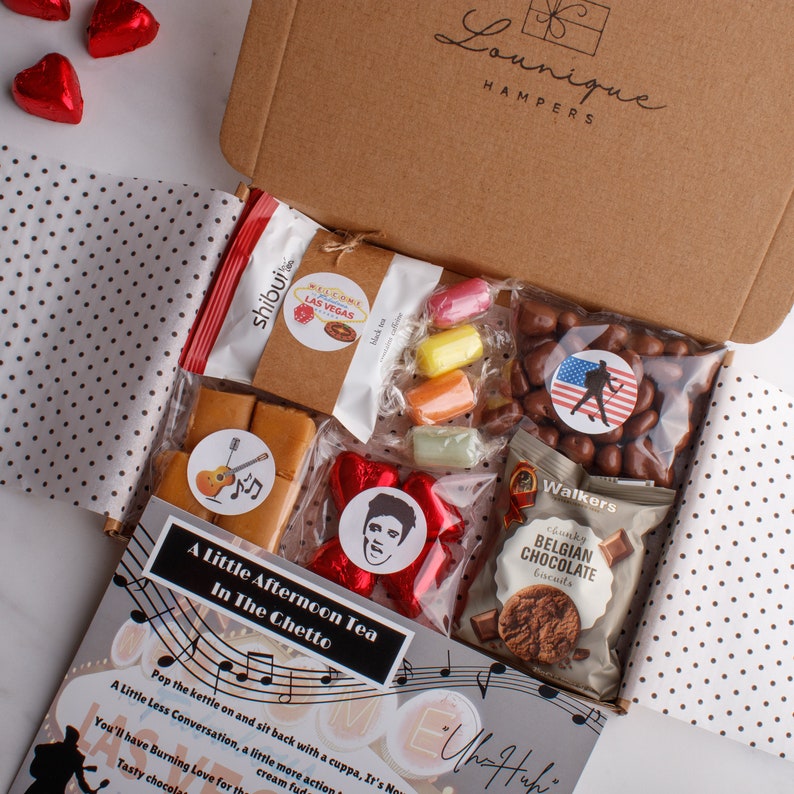 May include: A brown cardboard box filled with a variety of treats, including chocolate, sweets, and fudge. The box is decorated with a music note theme and the text "A Little Afternoon Tea In The Chetto".