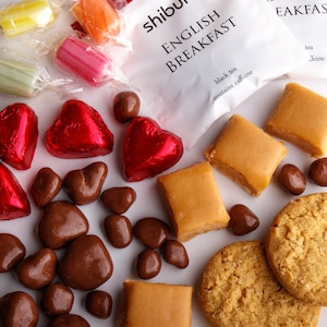 May include: A selection of sweets and treats, including chocolate hearts, chocolate sweets, caramel squares, shortbread biscuits, and wrapped sweets. The sweets are arranged on a white surface. The image also includes a tea bag packet with the text "English Breakfast black tea contains caffeine".