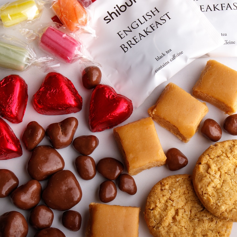May include: A selection of sweets and treats, including chocolate hearts, chocolate sweets, caramel squares, shortbread biscuits, and wrapped sweets. The sweets are arranged on a white surface. The image also includes a tea bag packet with the text "English Breakfast black tea contains caffeine".