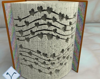 May include: An open book with pages carved to reveal a heart design. The book has a brown cover and the text "PATTERNS FOR 28 BOOK SIZES 16 IN CM + 12 IN INCHES" is visible. The book is a craft project.
