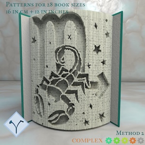 Könnte beinhalten: Ein aufgeschlagenes Buch mit einem detaillierten Skorpion-Design, das in die Seiten geschnitten wurde, umgeben von Sternen. Der Text oben lautet "PATTERNS FOR 28 BOOK SIZES 16 IN CM + 12 IN INCHES". Der Buchrücken ist grün.