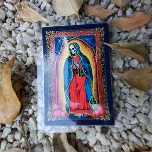 May include: A sticker featuring a colorful image of a skeleton figure wearing a blue and red robe with a gold halo. The figure is standing in front of a starry background with a gold frame.