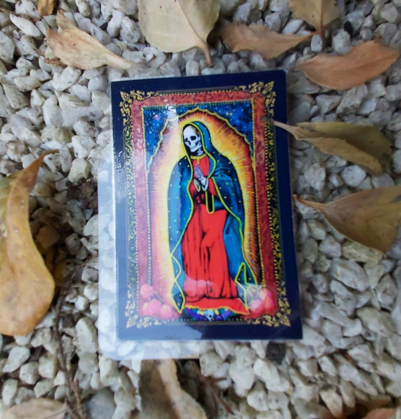 May include: A sticker featuring a colorful image of a skeleton figure wearing a blue and red robe with a gold halo. The figure is standing in front of a starry background with a gold frame.