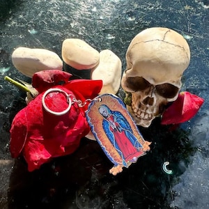 May include: A keychain featuring a colourful image of the Virgin Mary, a skull, and a red rose. The keychain is made of metal and has a silver ring.