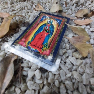 May include: A colorful sticker featuring a depiction of the Virgin Mary with a skull face. The sticker is on a background of white gravel.