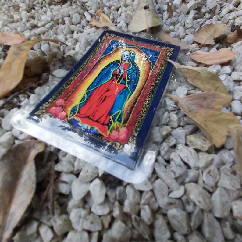 May include: A colorful sticker featuring a depiction of the Virgin Mary with a skull face. The sticker is on a background of white gravel.