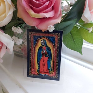May include: A colorful image of the Virgin Mary with a skeleton face, wearing a blue robe and red dress, against a starry background. The image is framed in gold and set against a dark blue background.