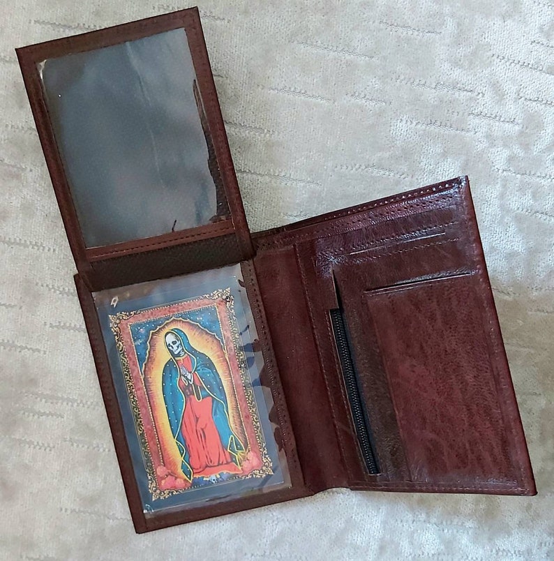 May include: A brown leather wallet with a clear plastic window insert. The window insert contains a colorful image of a woman wearing a blue and red robe. The wallet has a zippered pocket and a flap closure.