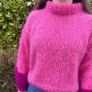 Peut inclure: Un pull rose vif &agrave; col montant et manches longues. Les manches ont des poignets magenta fonc&eacute; contrastants. Le pull est fait d'une mati&egrave;re douce et textur&eacute;e, parfait pour l'hiver.