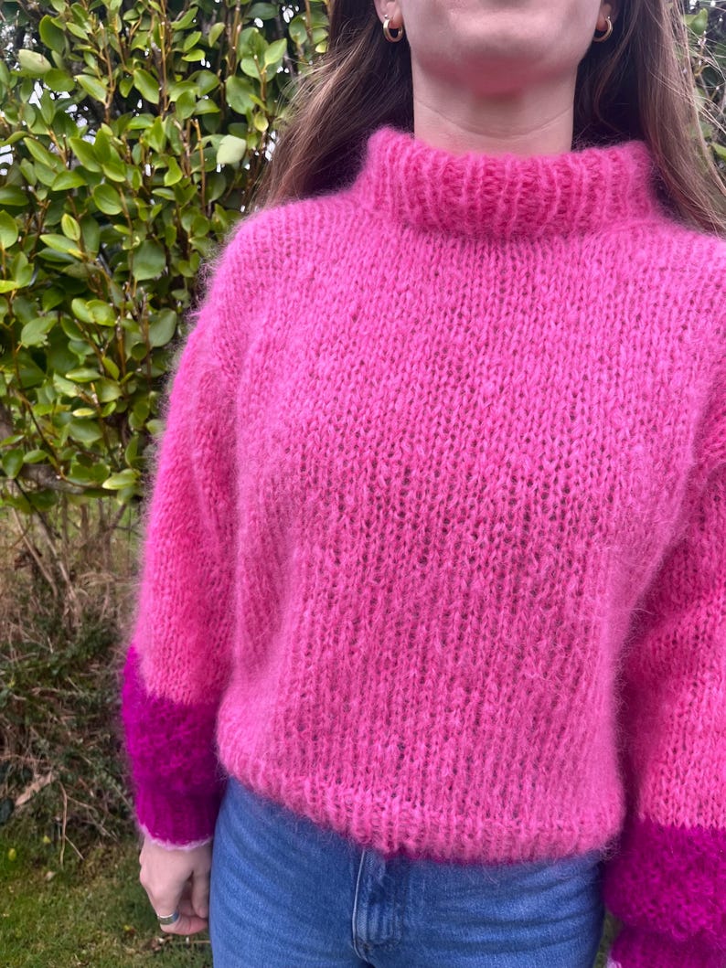 Peut inclure: Un pull rose vif &agrave; col montant et manches longues. Les manches ont des poignets magenta fonc&eacute; contrastants. Le pull est fait d'une mati&egrave;re douce et textur&eacute;e, parfait pour l'hiver.