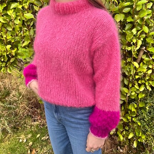 Peut inclure: Un pull rose vif tricot&eacute; &agrave; la main, avec un col montant et des manches longues. Les poignets sont d'une nuance de rose plus fonc&eacute;e. Le pull est fait d'une laine douce et duveteuse et est associ&eacute; &agrave; un jean bleu. Le fond est un buisson vert.