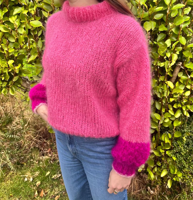 Peut inclure: Un pull rose vif tricot&eacute; &agrave; la main, avec un col montant et des manches longues. Les poignets sont d'une nuance de rose plus fonc&eacute;e. Le pull est fait d'une laine douce et duveteuse et est associ&eacute; &agrave; un jean bleu. Le fond est un buisson vert.