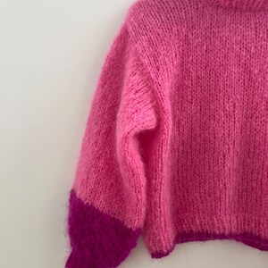 Peut inclure: Un pull rose vif tricot&eacute; &agrave; la main, avec un col montant et des manches longues. Les manches ont un poignet magenta fonc&eacute; contrastant. La texture douce et duveteuse de la laine est visible. Le pull est suspendu &agrave; un cintre en bois.