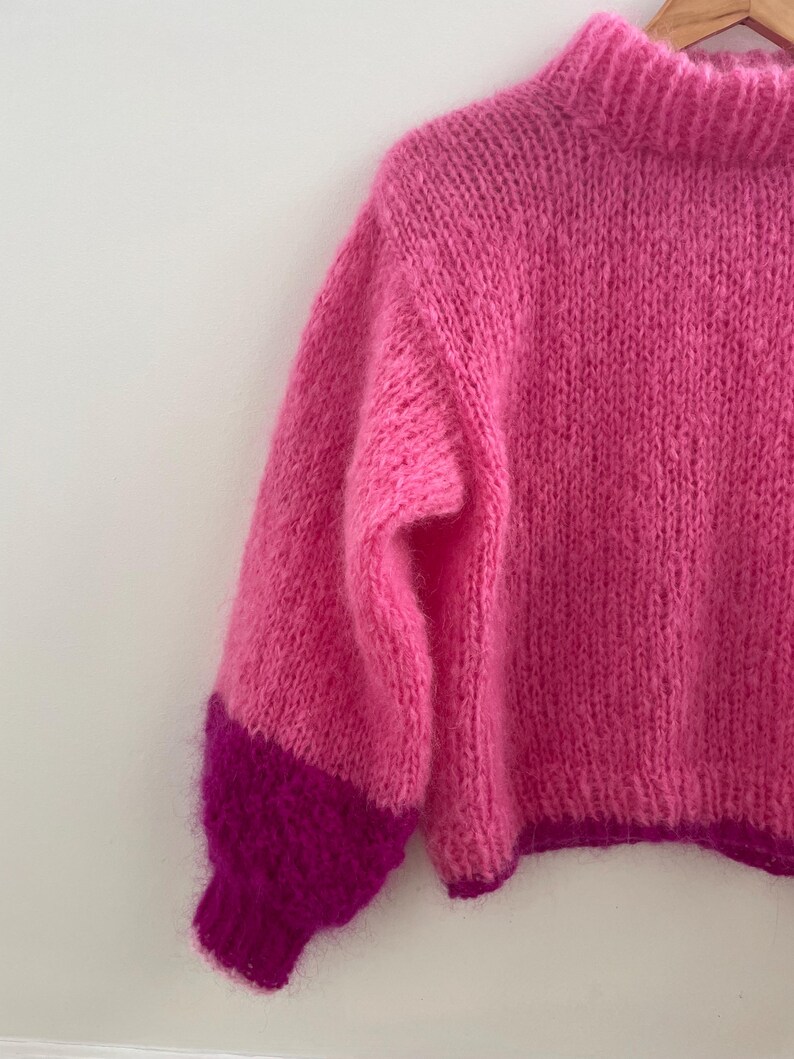 Peut inclure: Un pull rose vif tricot&eacute; &agrave; la main, avec un col montant et des manches longues. Les manches ont un poignet magenta fonc&eacute; contrastant. La texture douce et duveteuse de la laine est visible. Le pull est suspendu &agrave; un cintre en bois.