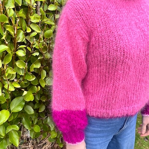 Peut inclure: Un pull rose vif &agrave; col montant et manches longues. Les poignets sont magenta fonc&eacute;. Le pull est fait d'une mati&egrave;re douce et duveteuse et est port&eacute; avec un jean bleu. Le fond pr&eacute;sente un feuillage vert.