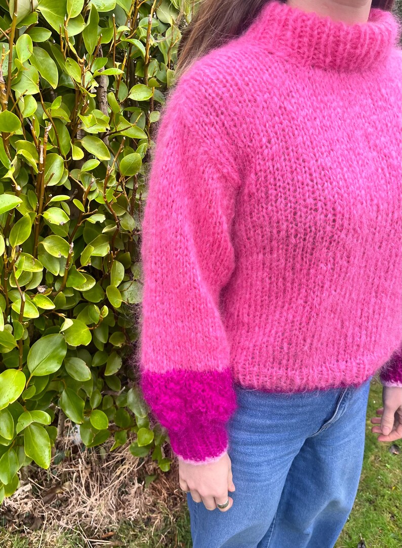 Peut inclure: Un pull rose vif &agrave; col montant et manches longues. Les poignets sont magenta fonc&eacute;. Le pull est fait d'une mati&egrave;re douce et duveteuse et est port&eacute; avec un jean bleu. Le fond pr&eacute;sente un feuillage vert.