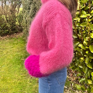 Peut inclure: Un pull court rose vif et duveteux avec un col montant et des manches longues. Les poignets sont d'une nuance de rose plus fonc&eacute;e. Le pull est associ&eacute; &agrave; un jean bleu. L'arri&egrave;re-plan pr&eacute;sente un feuillage et de l'herbe verts.