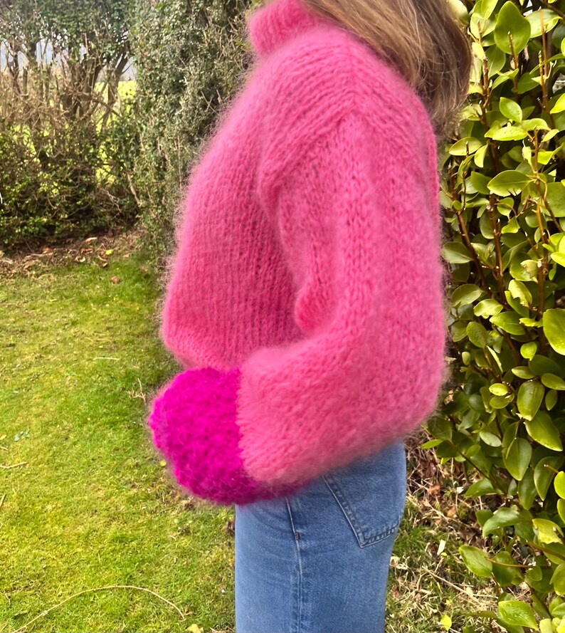 Peut inclure: Un pull court rose vif et duveteux avec un col montant et des manches longues. Les poignets sont d'une nuance de rose plus fonc&eacute;e. Le pull est associ&eacute; &agrave; un jean bleu. L'arri&egrave;re-plan pr&eacute;sente un feuillage et de l'herbe verts.