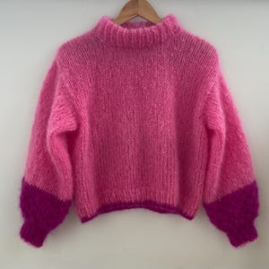 Peut inclure: Un pull rose tricot&eacute; &agrave; la main avec un col montant et des manches longues. Les manches ont des poignets magenta fonc&eacute;. Le pull est suspendu &agrave; un cintre en bois sur un fond blanc. La laine semble douce et duveteuse.