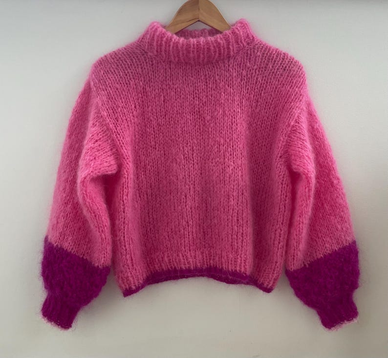 Peut inclure: Un pull rose tricot&eacute; &agrave; la main avec un col montant et des manches longues. Les manches ont des poignets magenta fonc&eacute;. Le pull est suspendu &agrave; un cintre en bois sur un fond blanc. La laine semble douce et duveteuse.