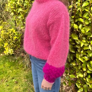 Peut inclure: Un pull rose vif tricot&eacute; &agrave; la main, avec un col montant et des manches longues. Les manches ont un poignet rose plus fonc&eacute;. Le pull est port&eacute; avec un jean bleu. Le fond est compos&eacute; de feuillage vert.