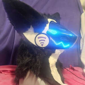 May include: A black and white furry head with large, pointed ears. The head features a glowing blue visor with a wave pattern and a white circular emblem with a purple music symbol. The head is resting on a gray base.