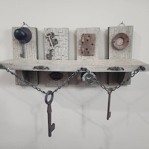 May include: A rustic wooden shelf with four decorative panels, each featuring vintage hardware like keys, a doorknob, and a hinge. The shelf is adorned with metal chains and hanging keys, creating a unique, antique-inspired display.