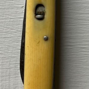 May include: A vintage pocket knife with a cream-colored handle and a partially visible blade. The handle has a rectangular shape with metal rivets and a metal clasp. The knife is closed and appears to be well-worn.