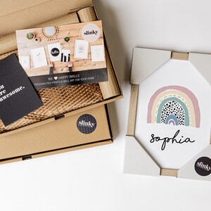 May include: A framed print with a rainbow design and black polka dots. The rainbow is in shades of pink, yellow, green, and blue. The name "Sophia" is written in black below the rainbow. The print is packaged in a cardboard box with a sticker that says "slinky".