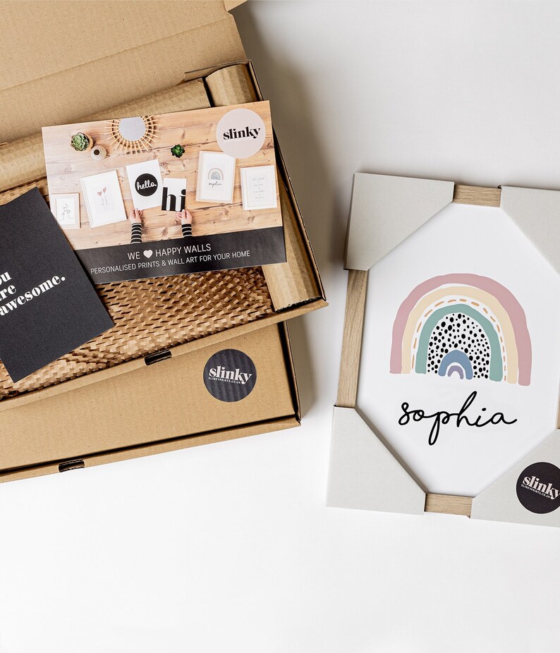May include: A framed print with a rainbow design and black polka dots. The rainbow is in shades of pink, yellow, green, and blue. The name "Sophia" is written in black below the rainbow. The print is packaged in a cardboard box with a sticker that says "slinky".