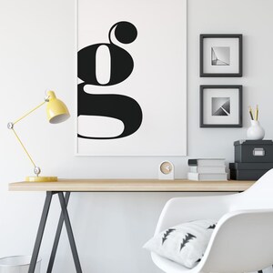 May include: A black and white poster with a large, stylized letter "G" on a white background. The poster is framed and hanging on a white wall above a wooden desk with a yellow lamp, a white chair, and a clock.