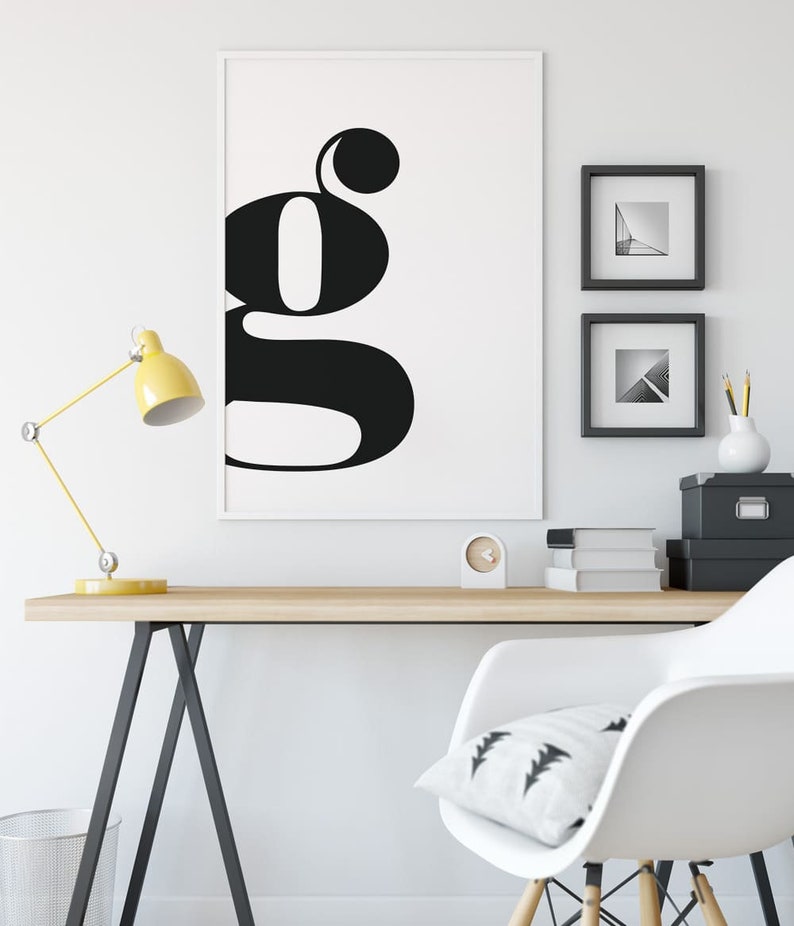 May include: A black and white poster with a large, stylized letter "G" on a white background. The poster is framed and hanging on a white wall above a wooden desk with a yellow lamp, a white chair, and a clock.