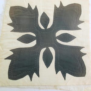 May include: A square, off-white textile with a dark gray, symmetrical floral design. The design is composed of stylized leaves and petal shapes, stitched with white thread. The edges of the textile are slightly frayed, giving it a vintage appearance.