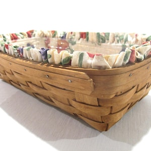 May include: A rectangular woven basket with a light brown finish. The basket's rim is lined with a ruffled fabric featuring a floral pattern in red, green, and purple. The basket is empty and sits on a white surface.