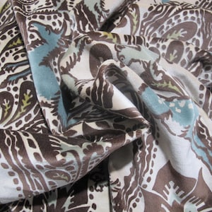 May include: Close-up of a fabric with a paisley pattern. The fabric features a repeating design in brown, light blue, and olive green on a cream background. The fabric is draped, showing folds and texture.