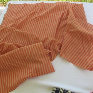 May include: Orange and cream-colored striped fabric, possibly ticking, is draped across a white surface. The fabric has a repeating pattern of thin red stripes. The material is folded and appears to be a length of cloth, possibly for sewing or crafting.