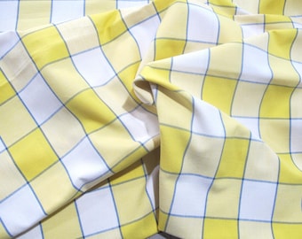 May include: Close-up of a fabric with a yellow, white, and blue plaid pattern. The fabric is folded and draped, showing the texture and design. The yellow squares are bordered by thin blue lines.