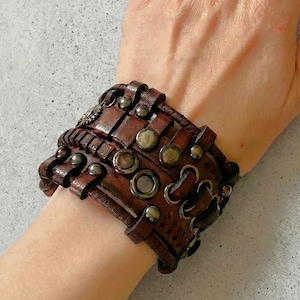 May include: A brown leather cuff bracelet with multiple straps and metal studs. The wide bracelet features decorative stitching and metal accents. The accessory is designed to be worn around the wrist.
