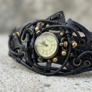 May include: A black leather steampunk-style wristwatch with a round, cream-colored watch face. The watch features Roman numerals and brass-colored accents. The intricate design includes leather straps and decorative metal elements.